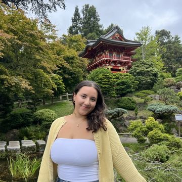 Sofia Pesantez standing in front a Japanese tea garden building, smiling at camera., wearing a yellow button up sweater, white top and jeans.